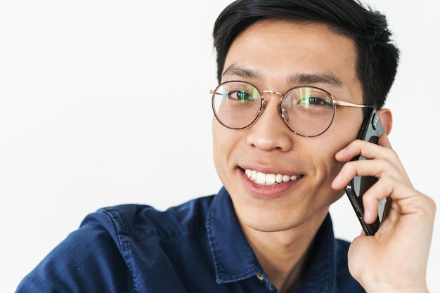 Foto van een brunette aziatische zakenman met een bril die in een stoel zit en op een smartphone praat terwijl hij op kantoor werkt, geïsoleerd over een witte muur