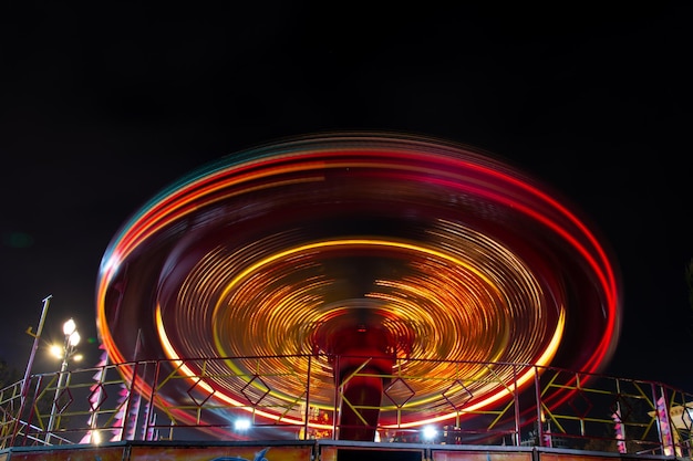 Foto van een carrousel bij nacht in een lange blootstelling in een pretpark. De gele en rodere tinten van de carrousellampen komen in beweging samen en geven het beeld een gloeiend schijfeffect.