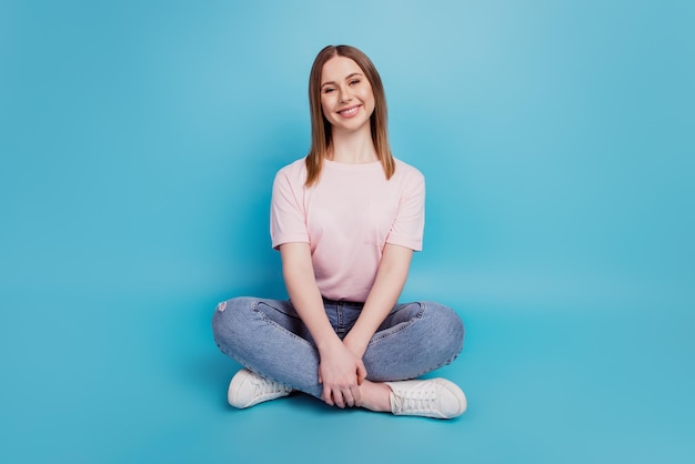 Foto van een charmante, vrij serene, kalme dame die op de vloer zit en een wit t-shirt draagt dat op een blauwe achtergrond wordt geïsoleerd