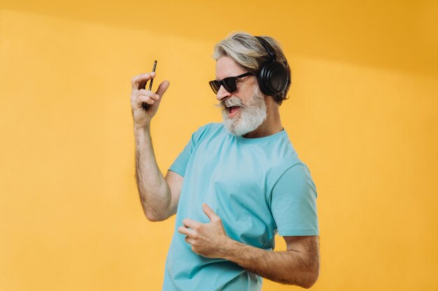 Foto van een expressieve grijsharige senior man in koptelefoon met een telefoon in zijn handen geïsoleerd op een gele achtergrond