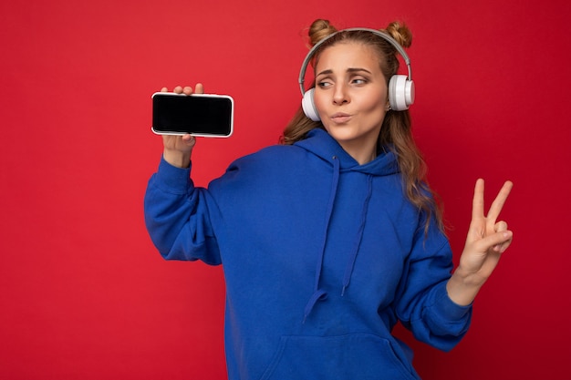 Foto van een fascinerende gelukkig lachende jonge vrouw die een stijlvolle blauwe hoodie draagt, geïsoleerd over rood