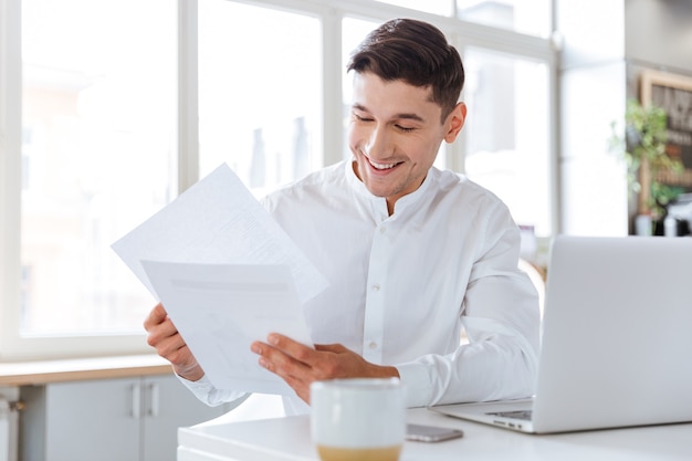 Foto van een gelukkige jonge man gekleed in een wit overhemd met documenten tijdens het gebruik van een laptopcomputer. Samenwerken. Documenten bekijken.