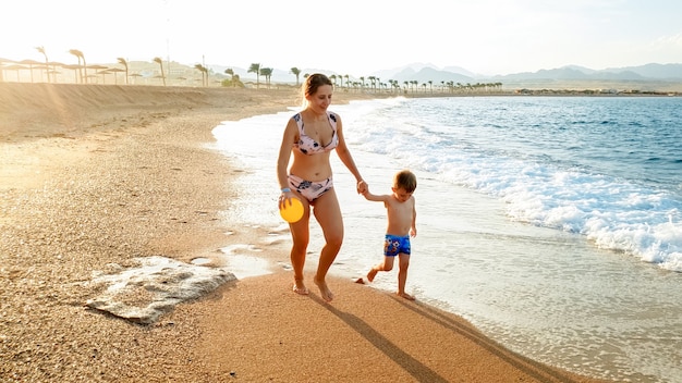 Foto van een gelukkige jonge moeder met een schattige 3 jaar oude peuterjongen die handen vasthoudt en op het zeestrand loopt tegen een prachtige zonsonderganghemel boven het wateroppervlak.