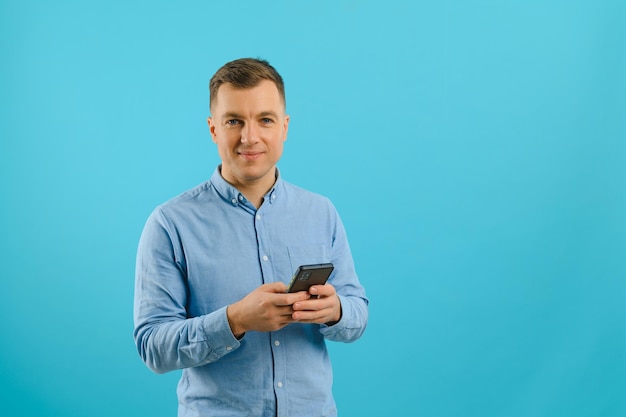 Foto van een gelukkige knappe man die een mobiele telefoon in de hand houdt, sms't. Jonge kerel met grote brede glimlach die zich voordeed op blauwe achtergrond.