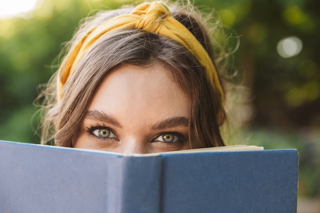 Foto van een gelukkige, mooie, mooie jonge vrouw in een groen park die een boek leest dat haar gezicht bedekt