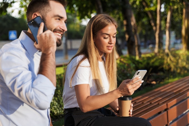 Foto van een gelukkige positieve jonge zakenman die via de mobiele telefoon praat terwijl zijn collega-vrouw aan het chatten in het natuurpark zit.