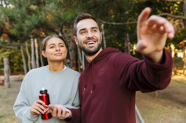 Foto van een gelukkige positieve sportvrouw en man die een mobiele telefoon buiten in een groen natuurpark gebruikt, wijzend opzij met een fles water.