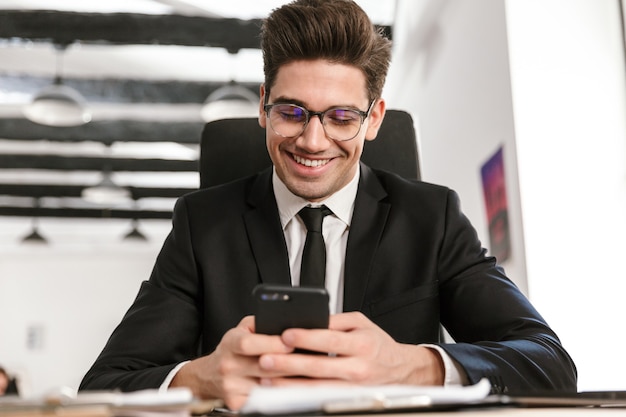 Foto van een gelukkige zakenman in een bril die op mobiele telefoon typt en glimlacht terwijl hij aan het bureau zit in een open kantoor