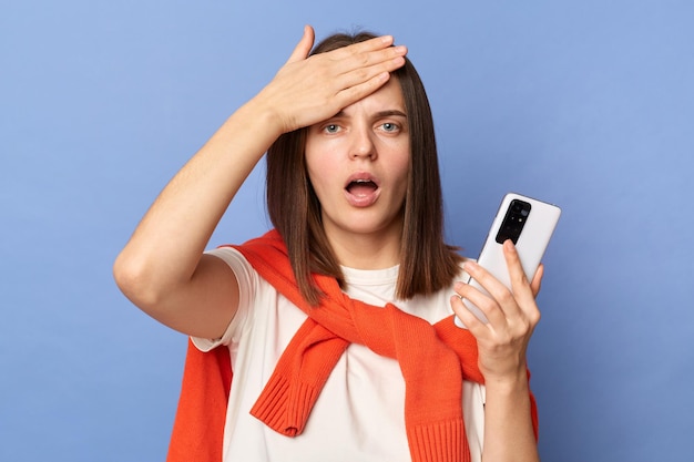 Foto van een geschokte jonge volwassen vrouw, gekleed in een wit T-shirt en een oranje trui die over de schouders is vastgebonden, geïsoleerd op een blauwe achtergrond terwijl ze haar mobiele telefoon vasthoudt en haar voorhoofd aanraakt en problemen heeft