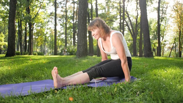 Foto van een glimlachende vrouw van middelbare leeftijd die yoga beoefent en mediteert in het park. Vrouw die zich uitstrekt en fitness doet op de mat in het bos