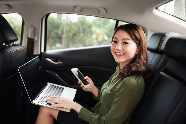 Foto van een jonge aziatische vrouw die aan de telefoon praat terwijl ze met een laptop in de auto werkt