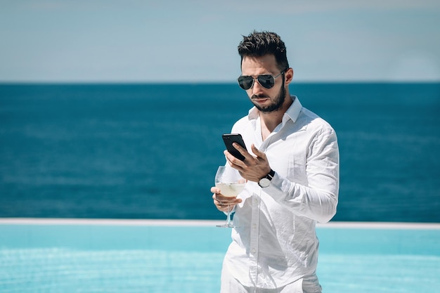 Foto van een jonge man in een wit overhemd die aan het zwembad staat en iets typt op de mobiele telefoon, mojito drinkt en geniet van een prachtig uitzicht op de blauwe zee. Reis- en bedrijfsconcept. Phuket. Thailand.