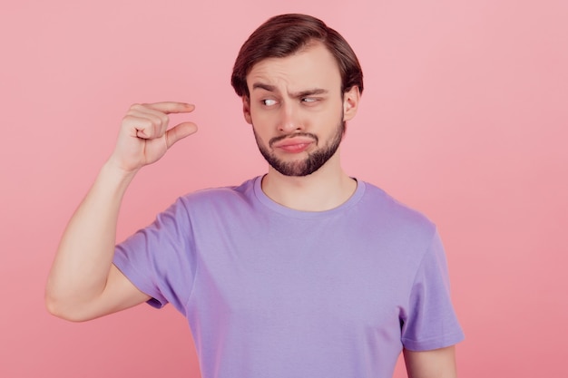 Foto van een jonge man, ongelukkig, verdrietig, van streek, toont kleine maatregelen van klein formaat met vingers die niet leuk zijn geïsoleerd over roze kleurachtergrond