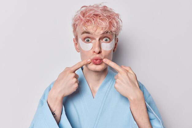 Foto foto van een jonge rozeharige man pruilt lippen kijkt verrassend naar de camera brengt schoonheidspleisters aan onder de ogen voor huidverjonging draagt een blauw gewaad verzorgt zichzelf geïsoleerd op witte achtergrond