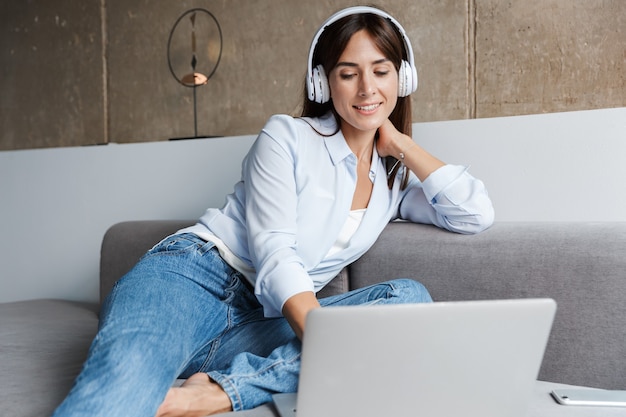 Foto van een jonge, vrolijke glimlachende vrouw die binnenshuis op de bank zit in de woonkamer en muziek luistert met een koptelefoon met behulp van een laptopcomputer.