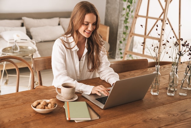 Foto van een jonge vrolijke positieve optimistische gembervrouw die binnenshuis op kantoor zit met een laptopcomputer.