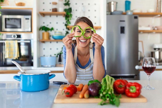 Foto van een jonge vrouw die lacht met plakjes groene paprika tijdens het koken van salade met verse groenten in het keukeninterieur thuis