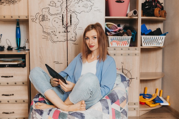 Foto foto van een jonge vrouw met een tablet die in een comfortabele zachte fauteuil zit, kenmerken van casual denimkleding.