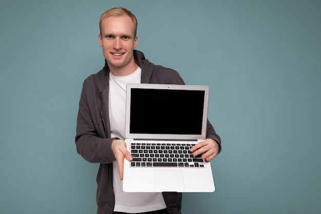 Foto van een knappe blonde man met een computerlaptop die naar een camera kijkt die over een blauwe muurachtergrond is geïsoleerd en een wit t-shirt en een grijze trui draagt