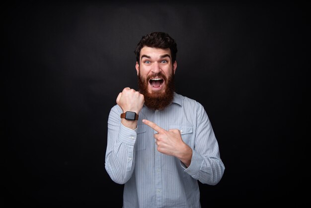 Foto van een knappe gefrustreerde man met baard, vertraging en wijzend op horloge, met geschokt gezicht, permanent over donkere geïsoleerde achtergrond