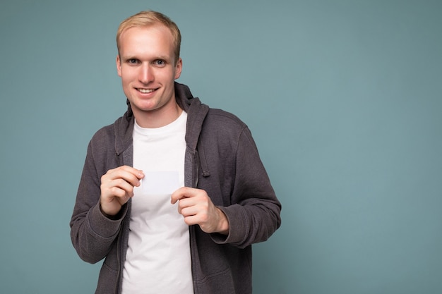 Foto van een knappe, gelukkige blonde man met een grijze trui en een wit t-shirt geïsoleerd over een blauwe achtergrondmuur met een creditcard die naar de camera kijkt