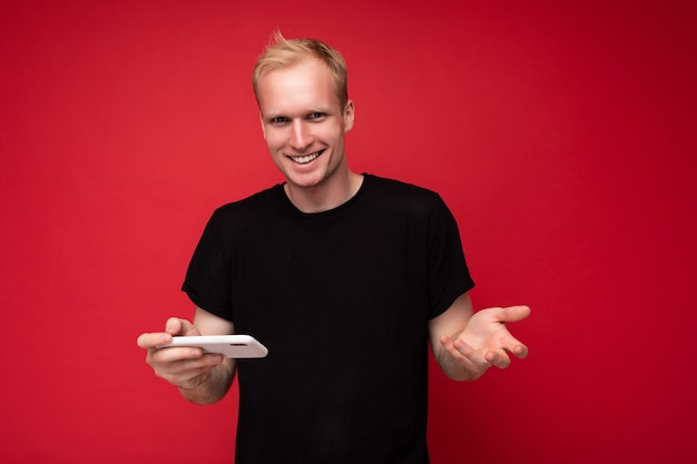 Foto van een knappe glimlachende positieve jonge blonde man geïsoleerd over een rode achtergrondmuur die een zwart t-shirt draagt en een mobiele telefoon gebruikt die sms schrijft en naar de camera kijkt.