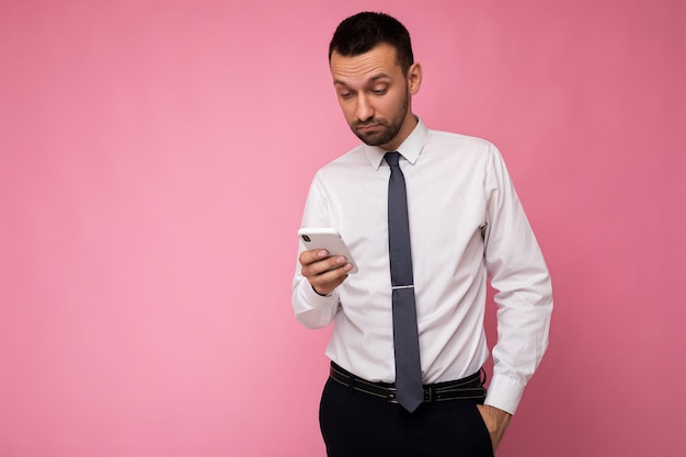 Foto van een knappe knappe man met een casual wit overhemd en stropdas geïsoleerd op een roze achtergrond met lege ruimte in de hand en met behulp van sms-berichten van mobiele telefoons die naar het gadjet-scherm kijken