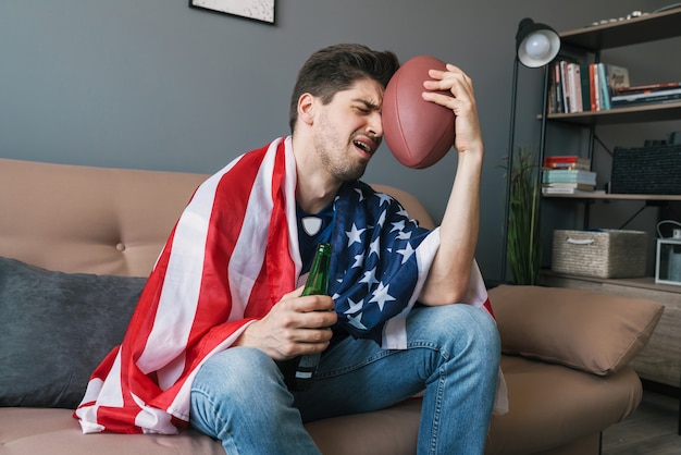 Foto van een knappe teleurgestelde man met een rugbybal en een Amerikaanse vlag die bier drinkt tijdens het kijken naar een sportwedstrijd in de woonkamer