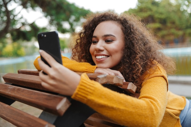 Foto van een lachende positieve mooie jonge krullende vrouw zit op een bankje in het park buiten met behulp van mobiele telefoon chatten.