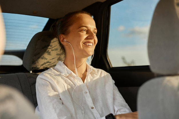 Foto van een lachende positieve vrolijke vrouw met een knotkapsel in een wit overhemd dat op de achterbank in de auto zit en glimlachend wegkijkt terwijl ze geniet van haar reis en muziek