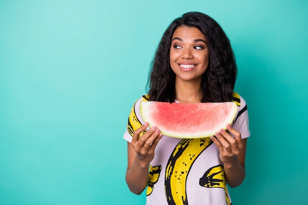 Foto van een meisje met een positieve donkere huid die een schijfje watermeloen vasthoudt, ziet er lege ruimte uit, geïsoleerd over een groenblauwe achtergrond