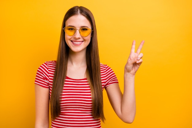 Foto van een mooi, vrij onzorgvuldig jong meisje lang kapsel glimlachend show v-teken gastvrije mensen student feestkleding zon specificaties gestreept wit rood shirt levendige gele kleur achtergrond