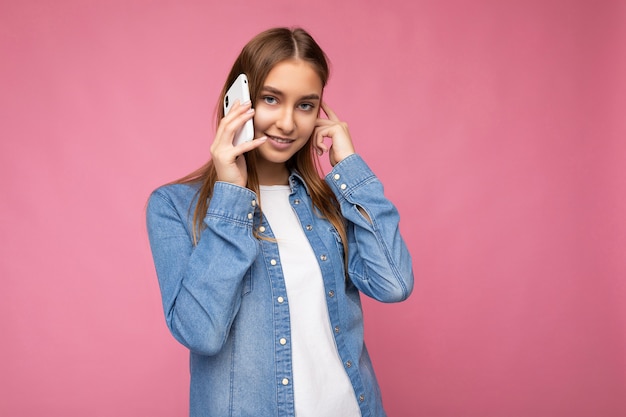 Foto van een mooie glimlachende gelukkige jonge blonde vrouwelijke peson die een casual blauw jean-shirt draagt, geïsoleerd over een roze achtergrond, in de hand houdt en praat op een mobiele telefoon die naar de camera kijkt.