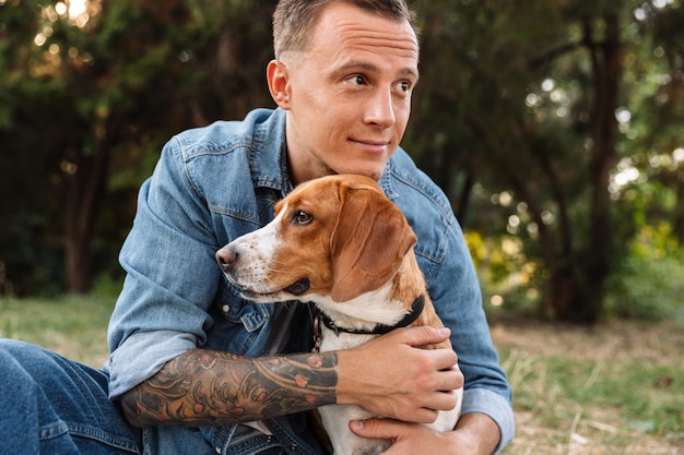 Foto van een mooie jonge man in spijkerkleding die lacht en zijn hond knuffelt in het park