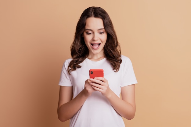 Foto van een opgewonden, vrolijk meisje houdt de telefoon vast met het scherm open mond poseren op een beige achtergrond