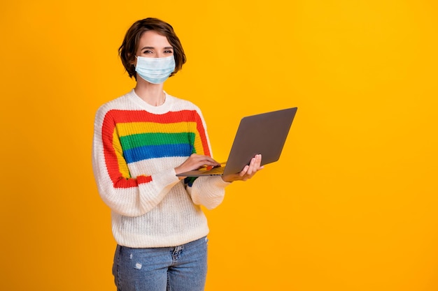 Foto van een positieve, aardige jonge vrouw die een laptop schrijft, draagt een medische respirator-sweater, een geïsoleerde gele achtergrond