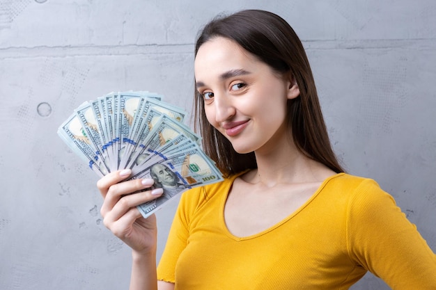 Foto van een rijke vrouw in eenvoudige kleren die een waaier van dollargeld houdt dat tegen een concrete muurachtergrond wordt geïsoleerd