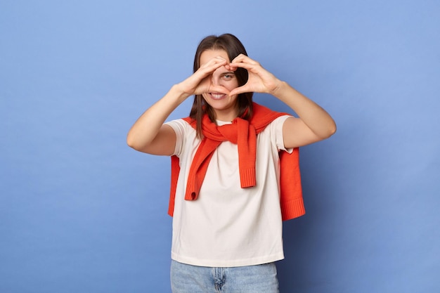 Foto foto van een romantische flirtende, vriendelijke jonge volwassen vrouw, gekleed in een wit t-shirt en een oranje trui die over de schouders is vastgebonden en hartvorm laat zien met de handen die tegen de blauwe muur staan