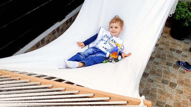 Foto van een schattige 3 jaar oude peuterjongen die ligt en ontspant in een witte hangmat in de achtertuin van het huis