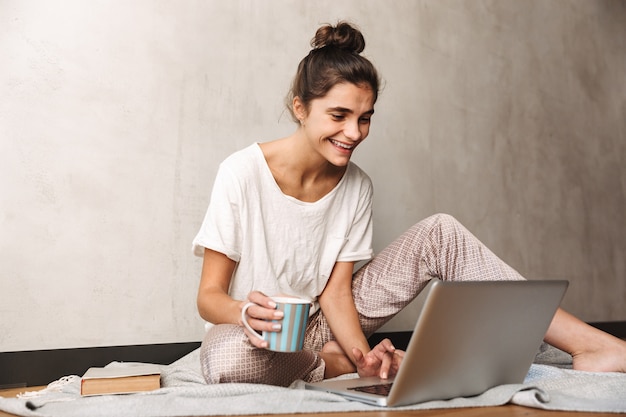 Foto van een schattige glimlachende vrouw die vrijetijdskleding draagt en koffie drinkt en typt op een laptop terwijl ze thuis op de vloer zit