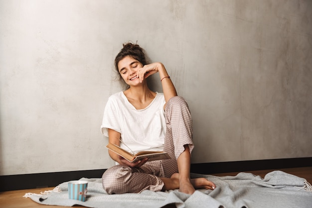 Foto van een schattige verbaasde vrouw die vrijetijdskleding draagt, koffie drinkt en een boek leest terwijl ze thuis op de vloer zit