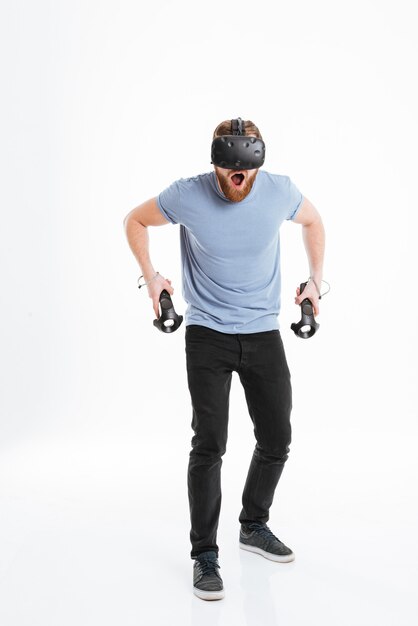 Foto van een schreeuwende jonge, bebaarde man met een virtual reality-apparaat terwijl hij joysticks in handen houdt.