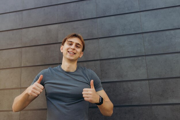 Foto van een sterke atleet die geïsoleerd staat over een grijze muurachtergrond Kijkend naar de camera die duim omhoog wijst, opgewonden vrolijke kerel