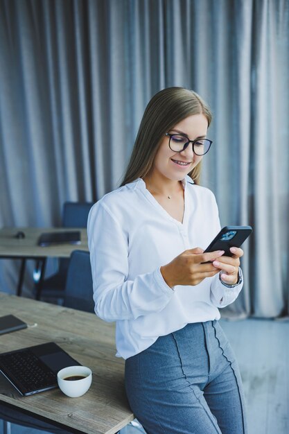 Foto van een succesvolle jonge vrouw in een shirt die lacht en op een laptop werkt terwijl ze telefoneert in een modern kantoor met grote ramen Werken op afstand met koffie