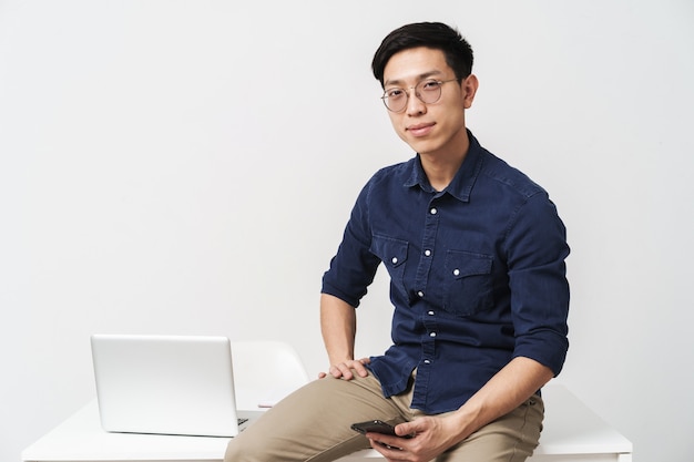 Foto van een tevreden aziatische zakenman met een bril die aan tafel zit en een smartphone vasthoudt terwijl hij met een laptop op kantoor werkt, geïsoleerd over een witte muur