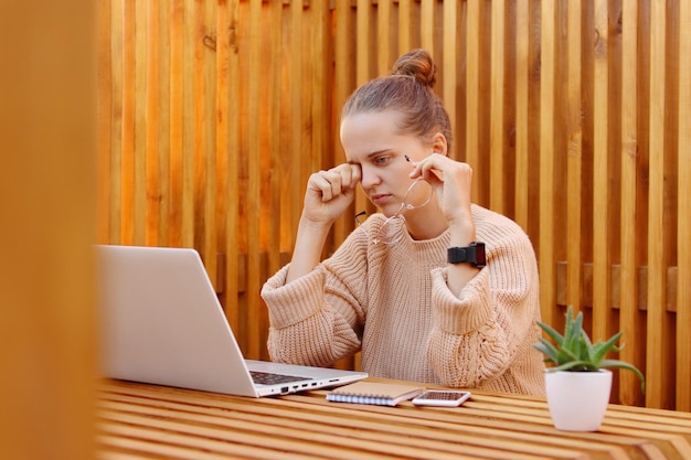 Foto van een uitgeputte vrouw met een beige trui die op kantoor aan een notitieboekje werkt tegen een houten muur die in haar ogen wrijft en moe is, verdrietig en overstuur is