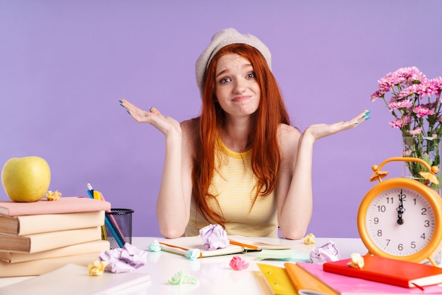 Foto foto van een verbaasd studentmeisje dat handen overgeeft terwijl ze aan het bureau zit met oefenboeken geïsoleerd over een paarse muur