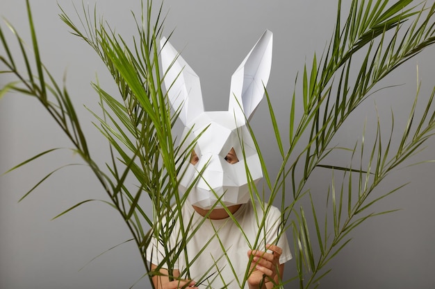 Foto van een verlegen onbekende vrouw met een wit overhemd en een konijnenmasker die zich voordeed op een grijze achtergrond die zich verschuilt achter groene palmtakken en wil niet opgemerkt worden