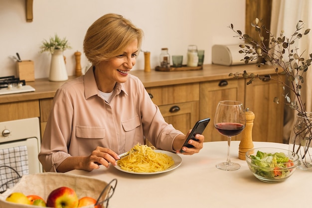 Foto van een volwassen positieve senior vrouw die thuis in de keuken zit met een mobiele telefoon.