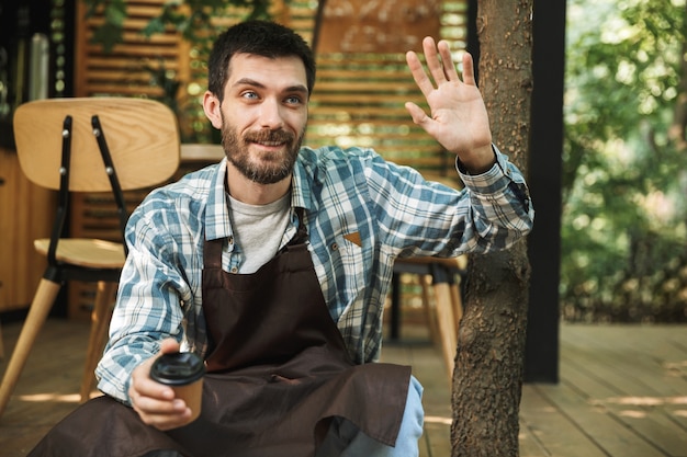 Foto van een vriendelijke ober met een schort die op een houten vloer zit terwijl hij in een café of koffiehuis buiten werkt